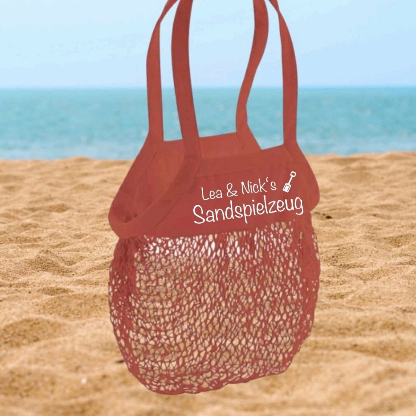 Personalisierte Sandspielzeugtasche in Rotbraun mit weißem Namensaufdruck am Strand