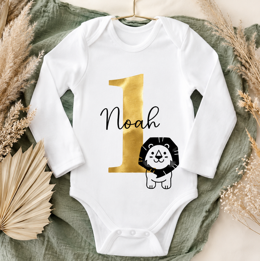 Weißer Babybody zum 1. Geburtstag mit goldener Zahl 1, personalisiertem Namen und niedlichem Löwenmotiv aus Bio-Baumwolle. Geschenk zum ersten Geburtstag für Jungen und Mädchen.