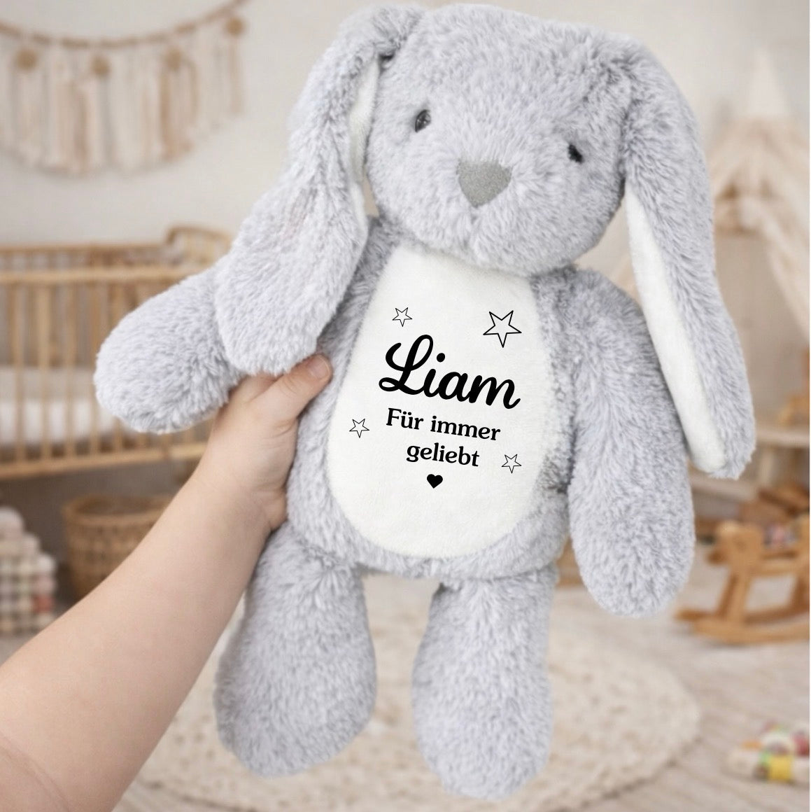 Ein weicher, grauer Plüschhase mit langen Schlappohren wird in einer Hand im Kinderzimmer gehalten. Auf dem weißen Bauch steht der Name Liam und der Spruch Für immer geliebt mit kleinen Sternen.