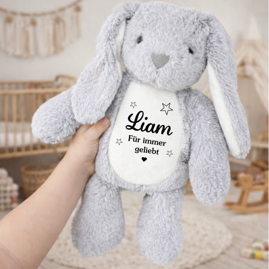 Ein weicher, grauer Plüschhase mit langen Schlappohren wird in einer Hand im Kinderzimmer gehalten. Auf dem weißen Bauch steht der Name Liam und der Spruch Für immer geliebt mit kleinen Sternen.